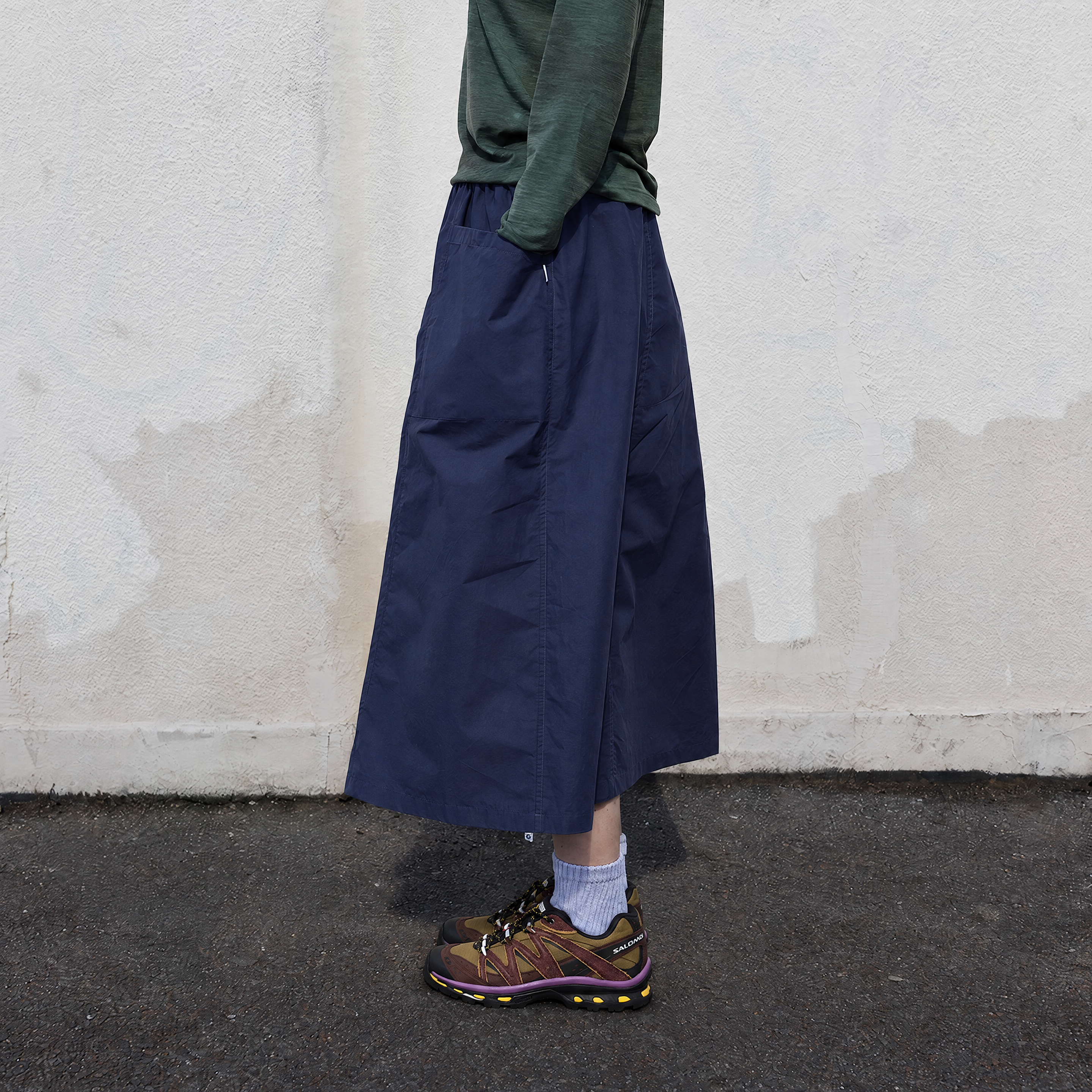 A person wearing a dark navy wide-leg midi skirt, a dark green long-sleeve top, and brown hiking shoes, standing against a textured off-white wall.
